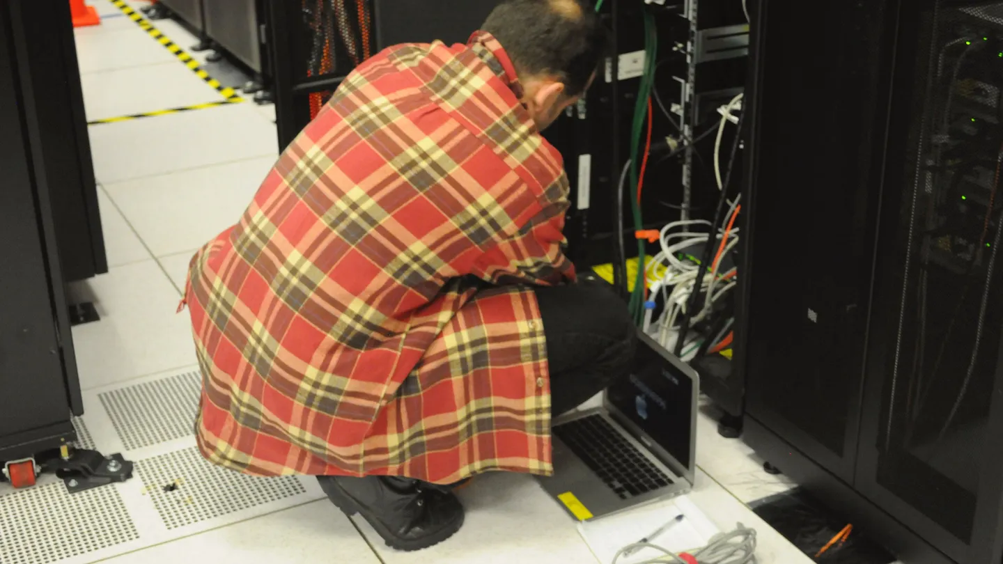 A technician crouching at an open server rack with a laptop in a data center